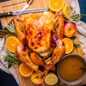 roasted whole turkey on a cutting board with bowl of gravy with fruit and herbs around.