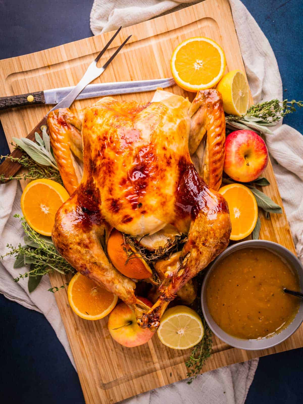 roasted whole turkey on a cutting board surrounded by fruit and fresh herbs and gravy bowl.