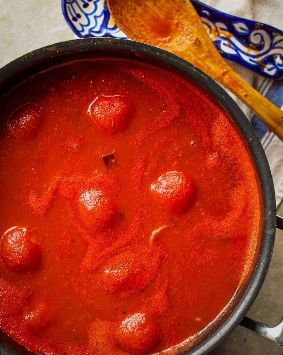 whole tomatoes simmering in a large pot of italian gravy with a wooden spoon next to it