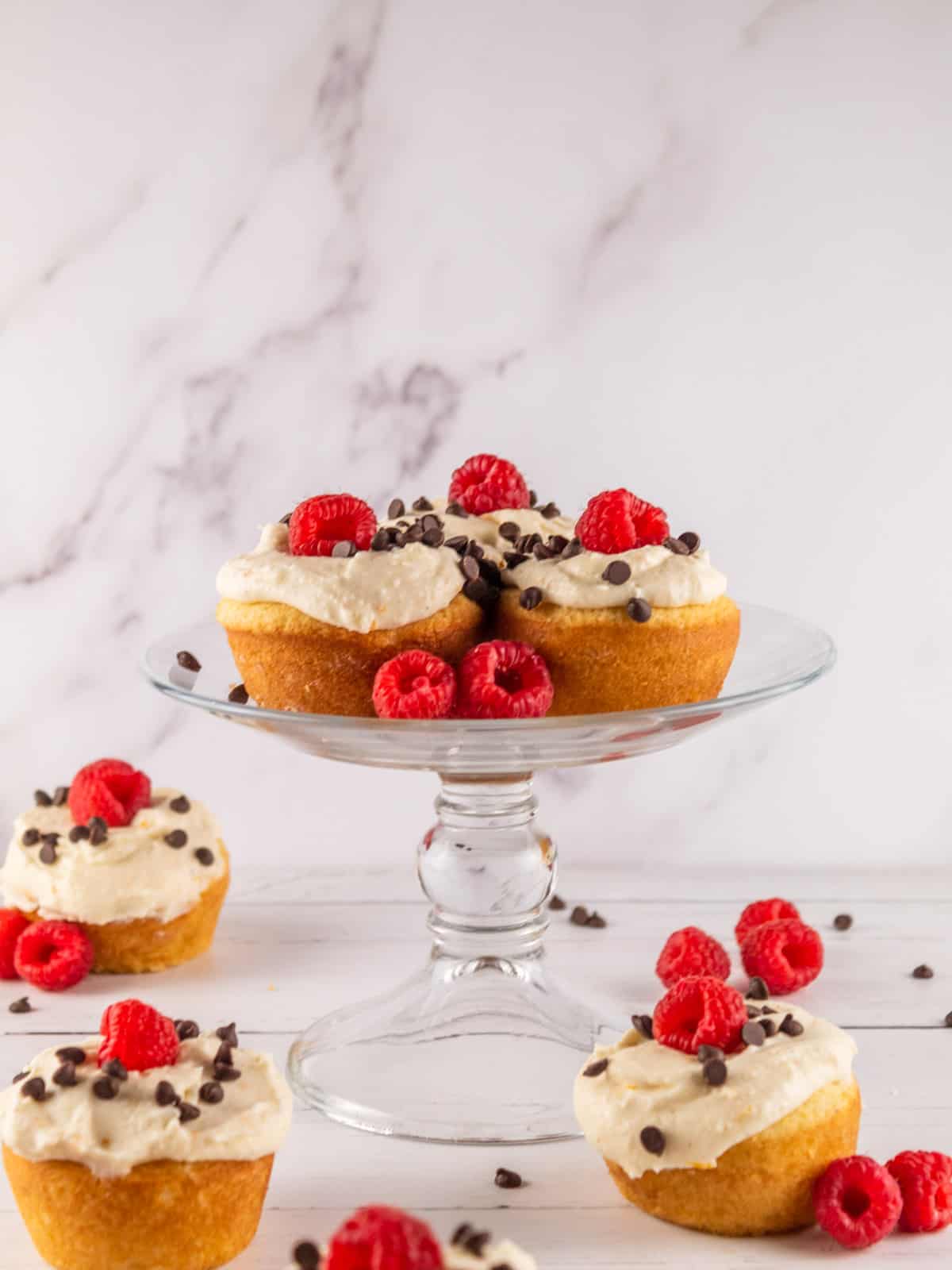 cupcakes on a clear glass stand with cannoli frosting, mini chocolate chips and fresh raspberries.