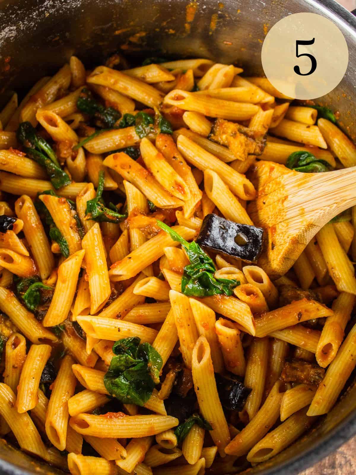 wooden spoon stirring penne pasta with eggplant, spinach and marinara sauce.