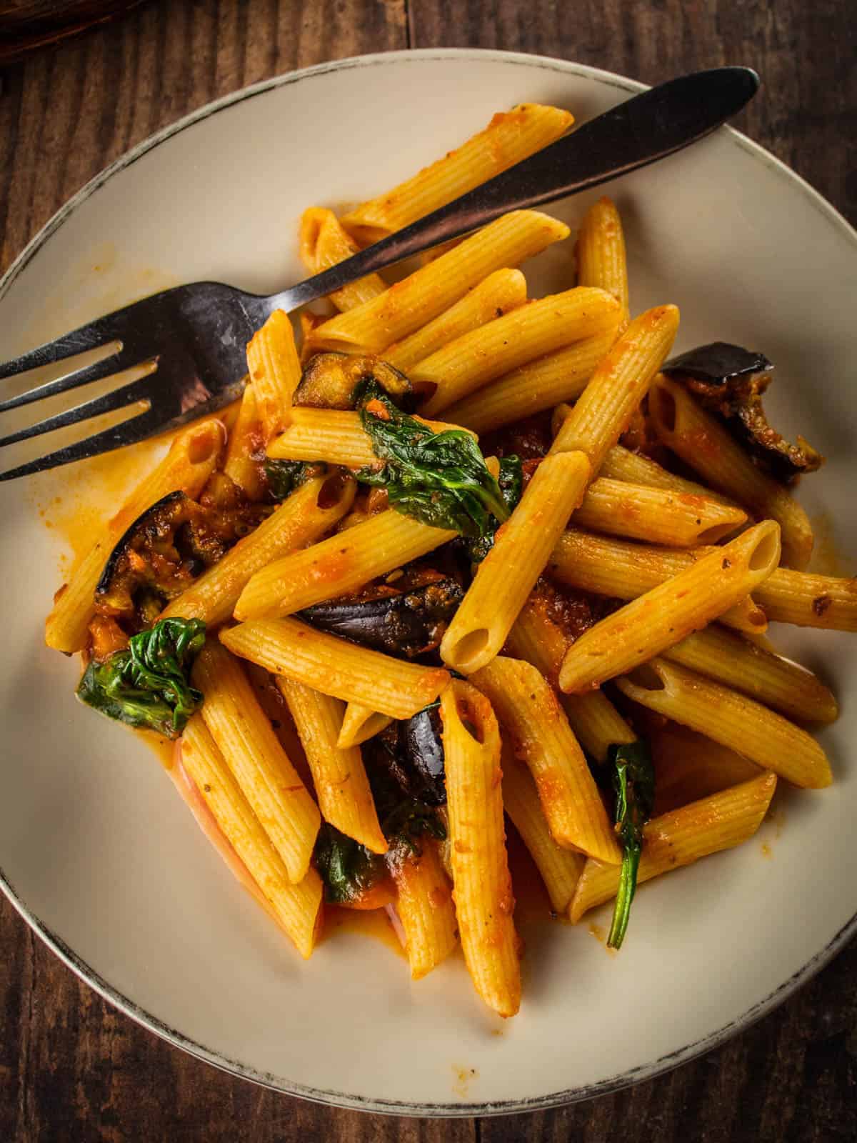 plate of penne pasta with roasted eggplant pieces, baby spinach and marinara sauce with a fork.