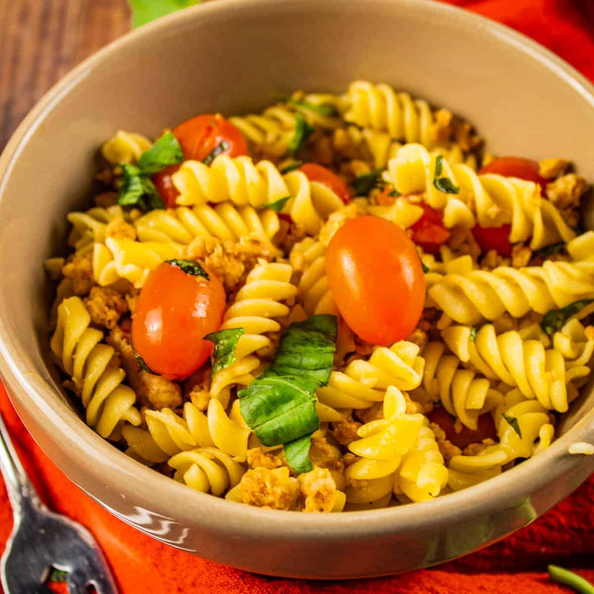 rotini pasta in a tan bowl with italian sausage, grape tomatoes and fresh basil