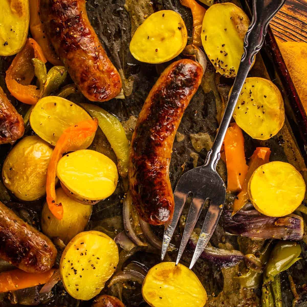 browned sausages on a sheet pan with halved potatoes, sliced peppers and a serving fork