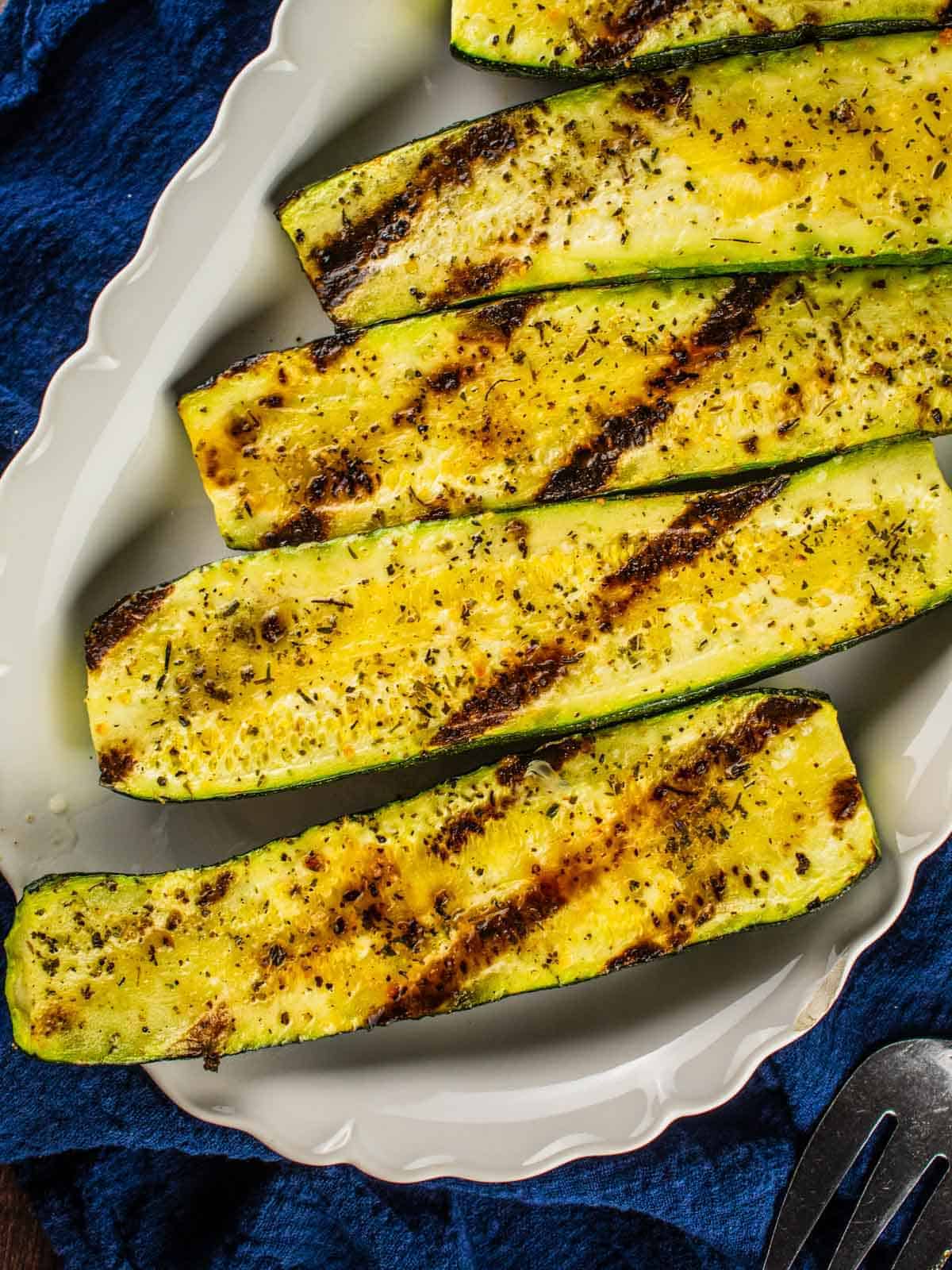 grilled seasoned zucchini halves on a white platter with a blue towel underneath it