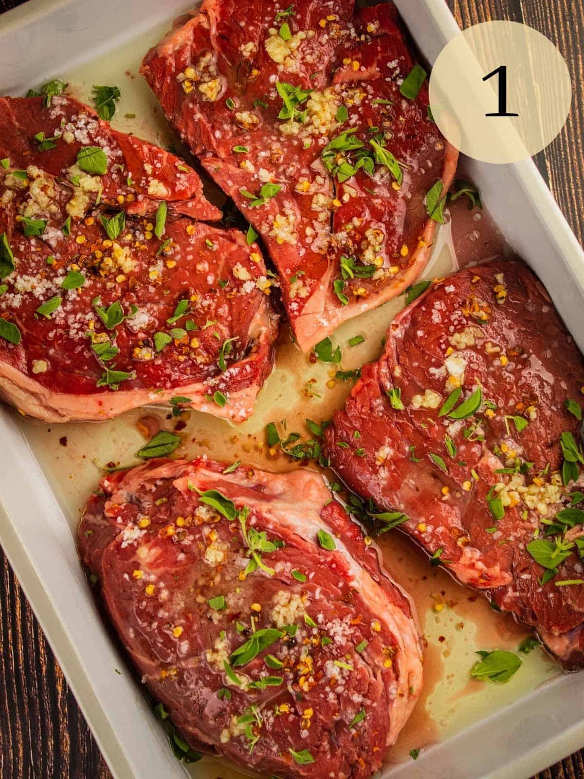 raw sirloin steaks marinating in oil, vinegar and seasoning in a white baking dish.