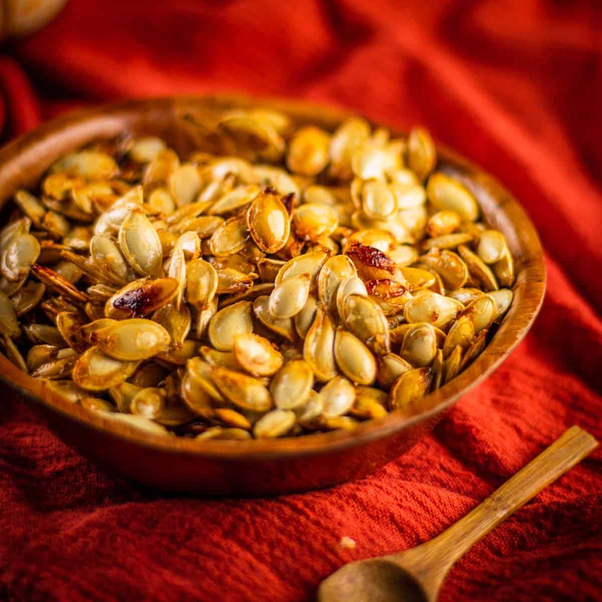 wooden bowl filled with roasted pumpkin seeds on a orange napkin with a small wooden spoon