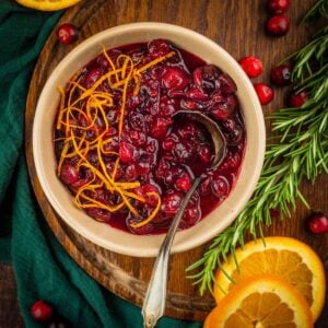 tan bowl filled with cranberry sauce topped with orange zest next to orange slices and fresh cranberries