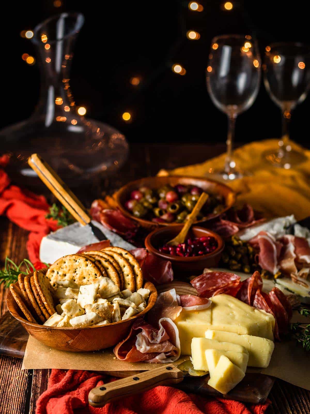 charcuterie board with meat, cheese and crackers with wine decanter, lights and wine glasses in the background