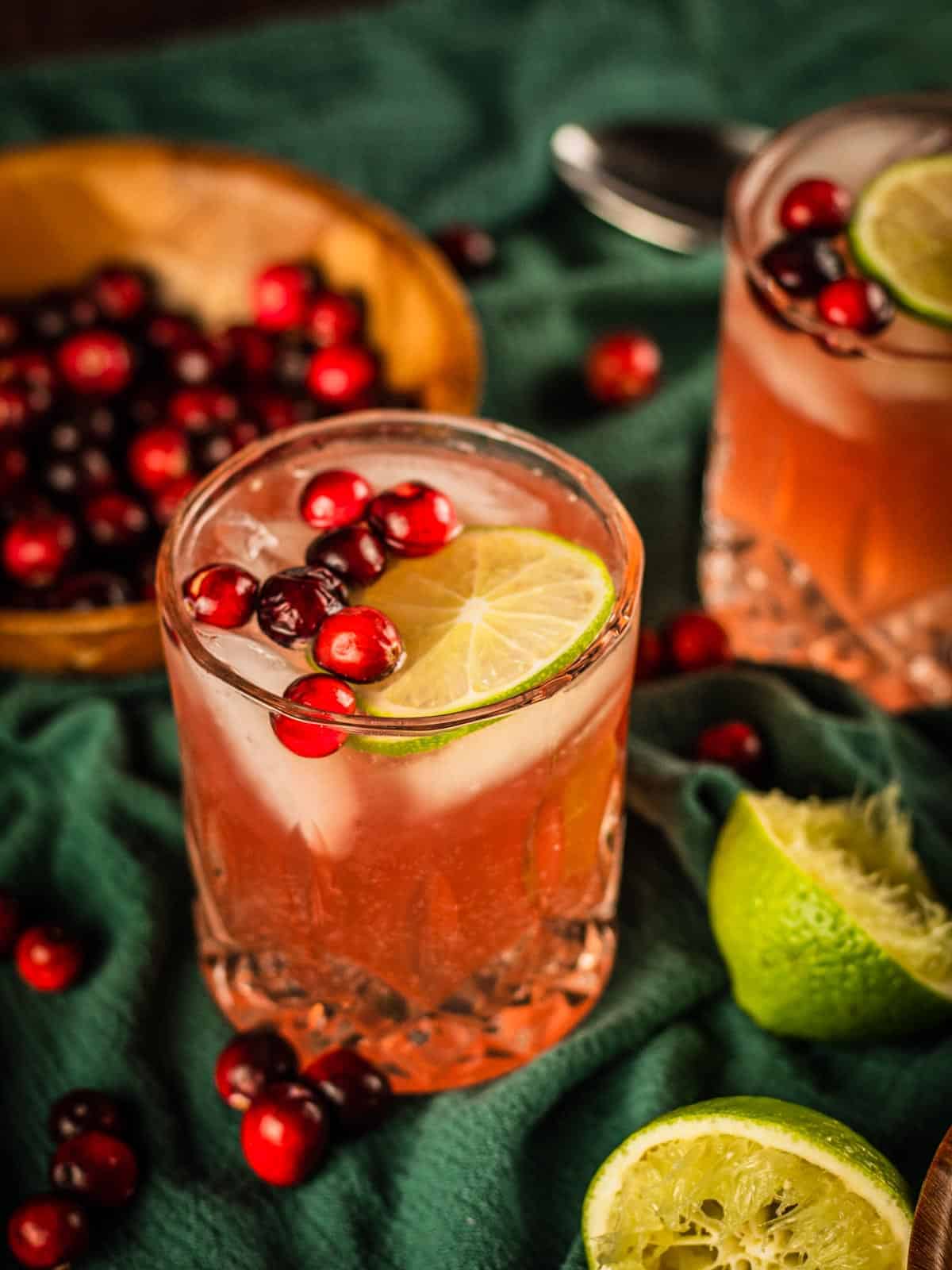two clear glasses filled with cranberry moscow mule cocktail, fresh cranberries and a lime wheel