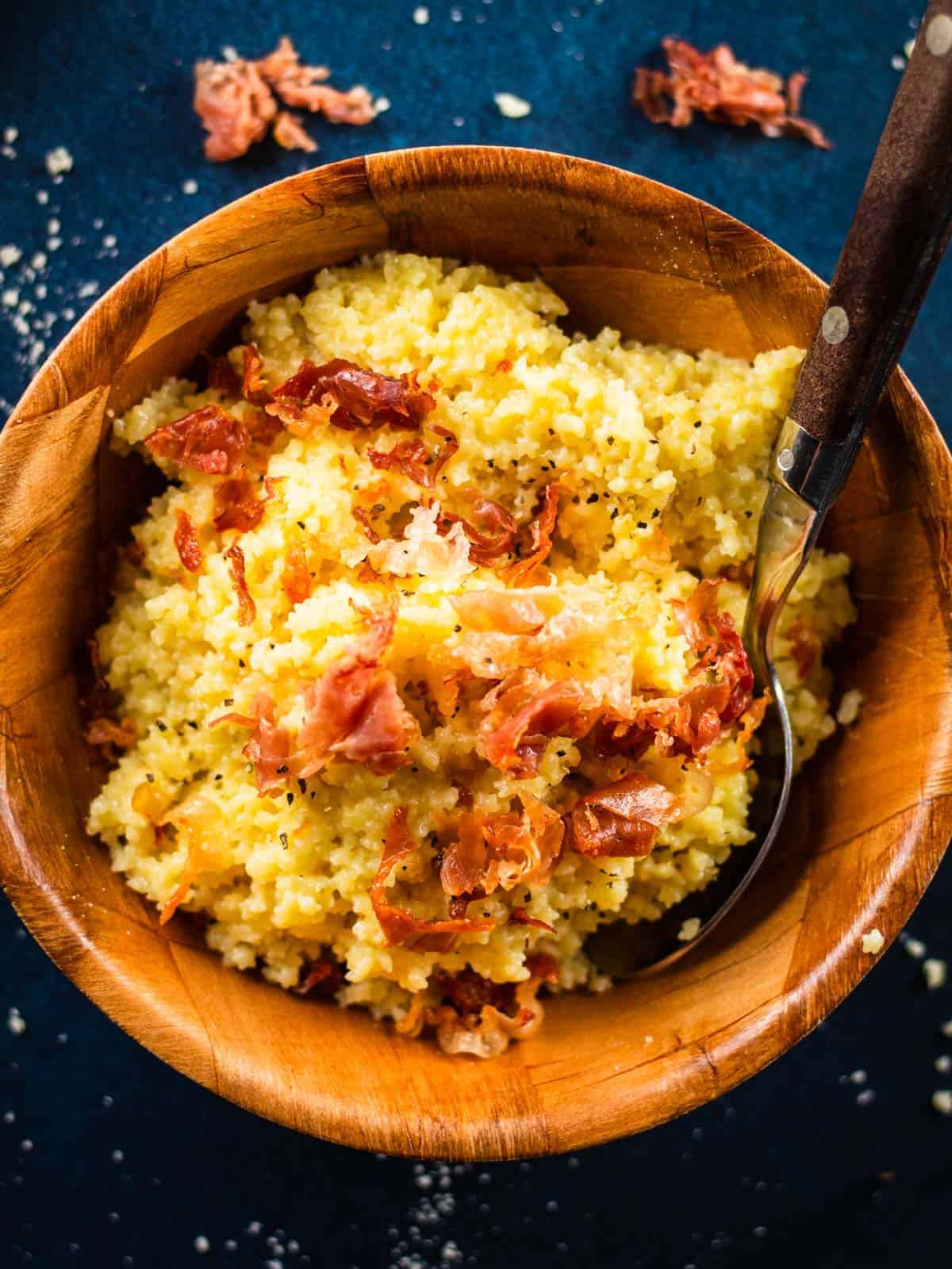 cheesy pastina pasta topped with crispy prosciutto in a wooden bowl with a spoon