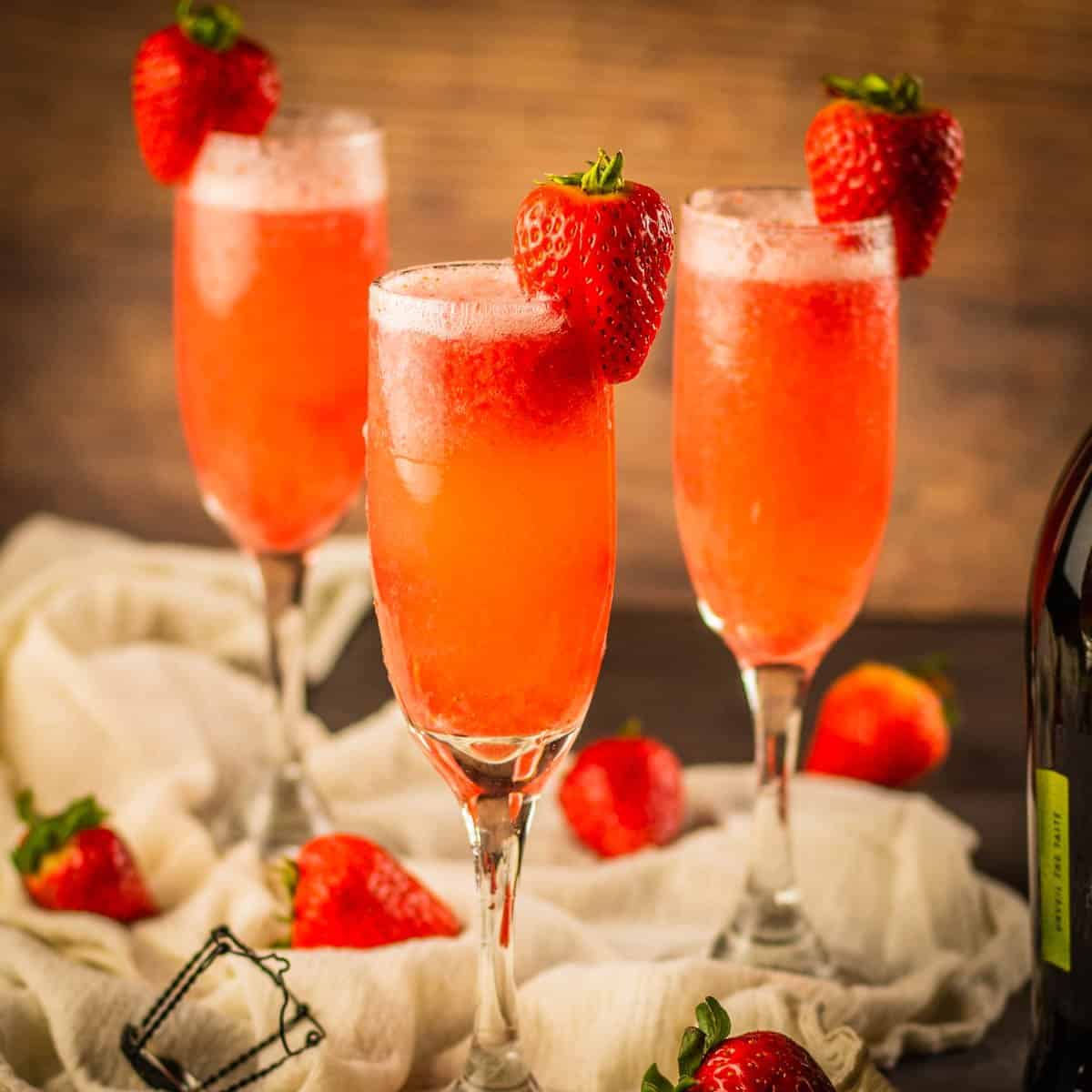 three champagne flutes filled with prosecco and strawberry puree and garnished with fresh strawberry