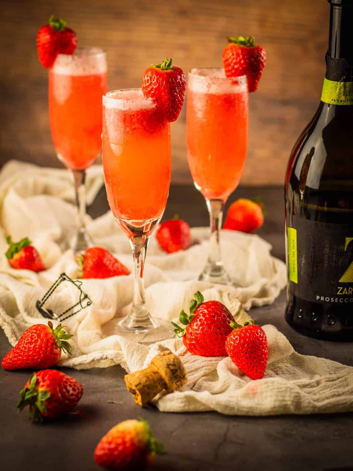 three champagne flutes with prosecco and strawberry puree garnished with a fresh strawberry