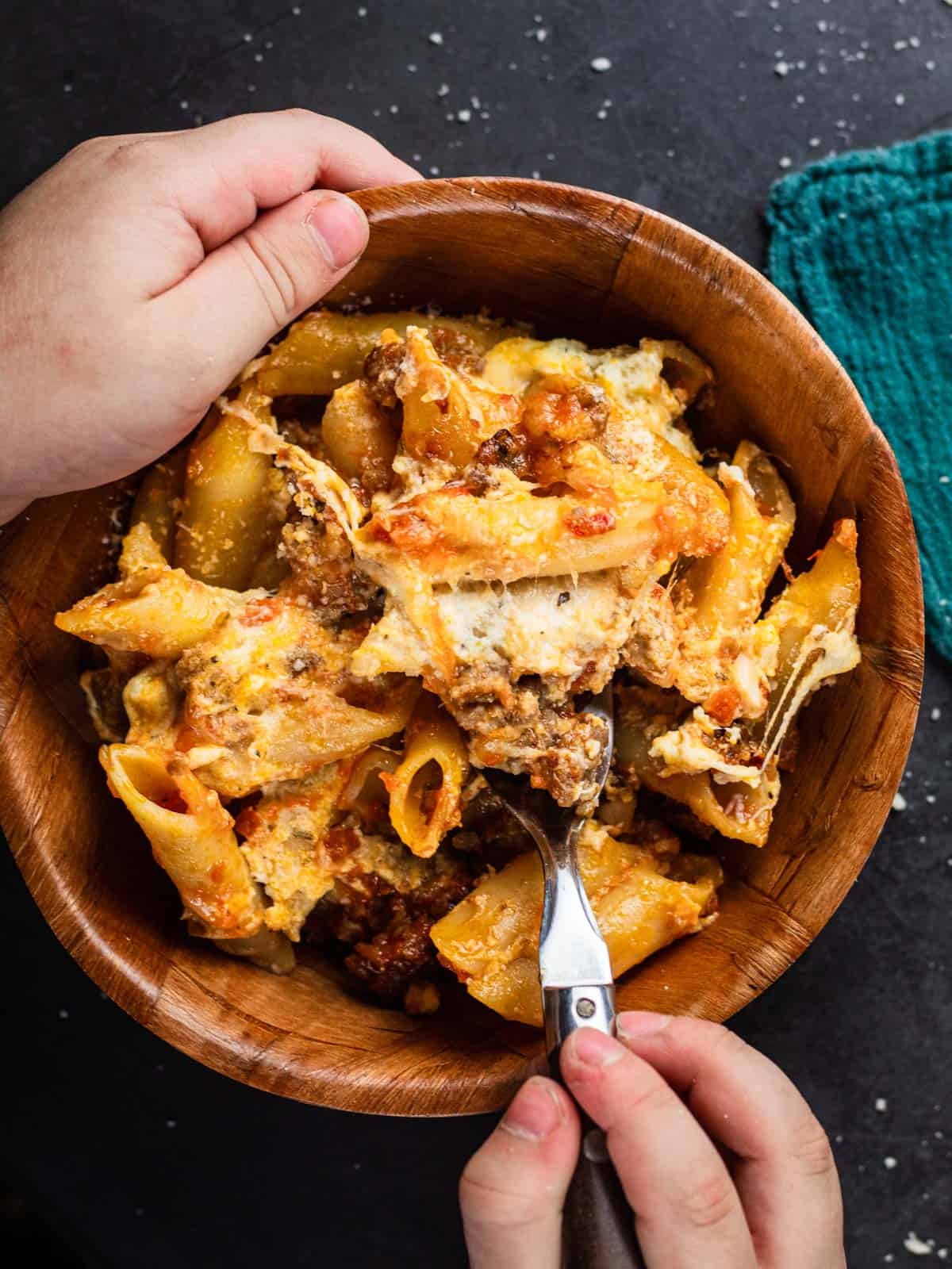 wooden bowl filled with cheesy mostaccioli with red sauce and a hand holding the bowl 