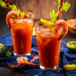 two bloody marys garnished with shrimp, bacon, celery, olives and peppers
