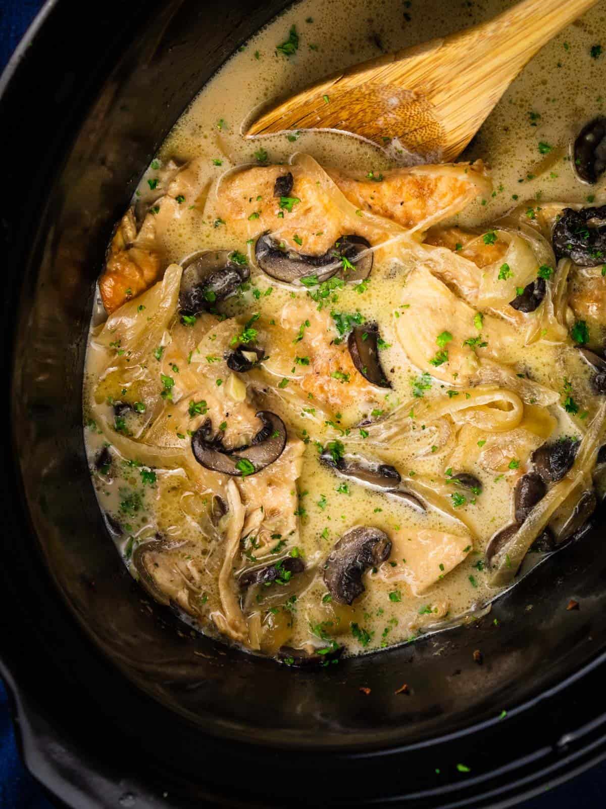 wooden spoon stirring creamy chicken marsala in a crock pot with onions and mushrooms