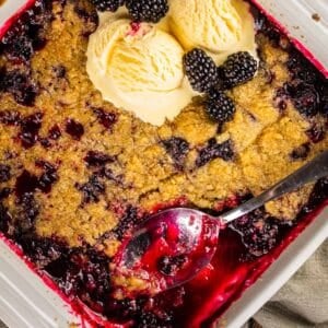 white square dish with blackberry crumble topped with ice cream and fresh blackberries.