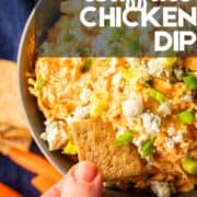 hand dipping cracker into buffalo chicken dip topped with green onion and blue cheese crumbles.