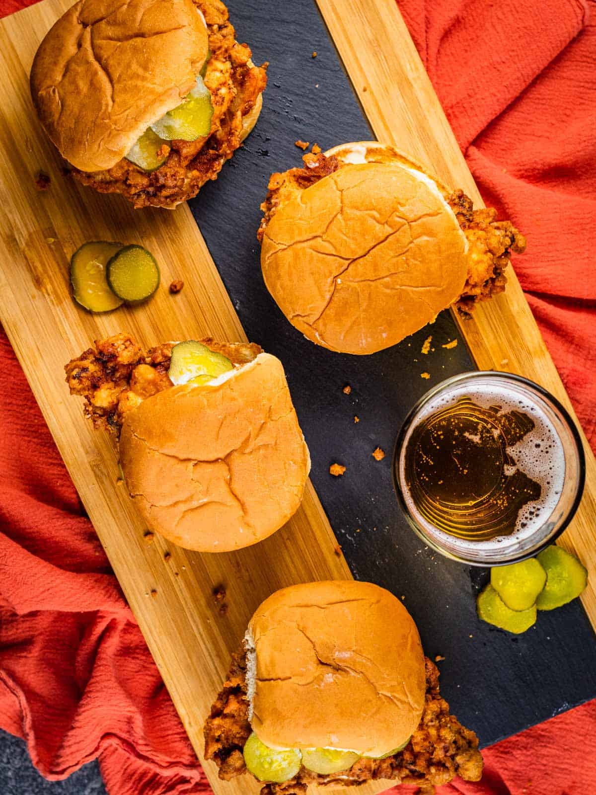 fried chicken sandwiches on a wooden tray with a beer and pickles laying around.