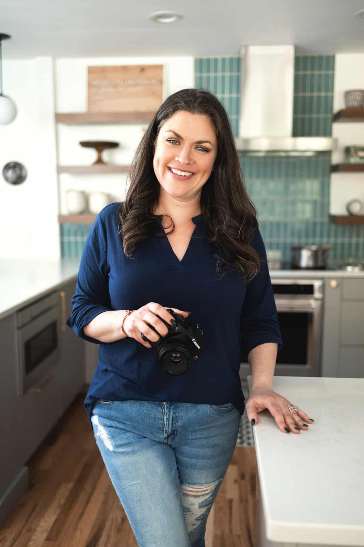 woman standing in a kitchen, holding a camera with the other hand on the counter.