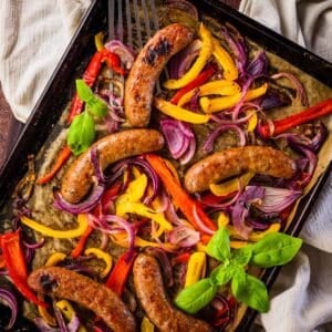 baked sausage, peppers and onions on a sheet pan with fresh basil sprigs around.