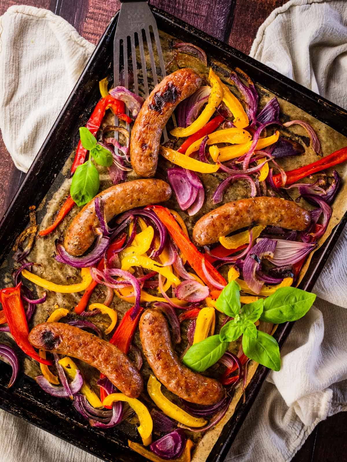 italian sausage, peppers and red onions baked on a sheet pan garnished with fresh basil sprigs.