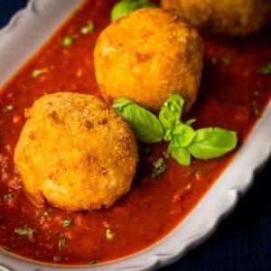 italian rice balls fried and set on marinara on a white platter with fresh basil sprigs.