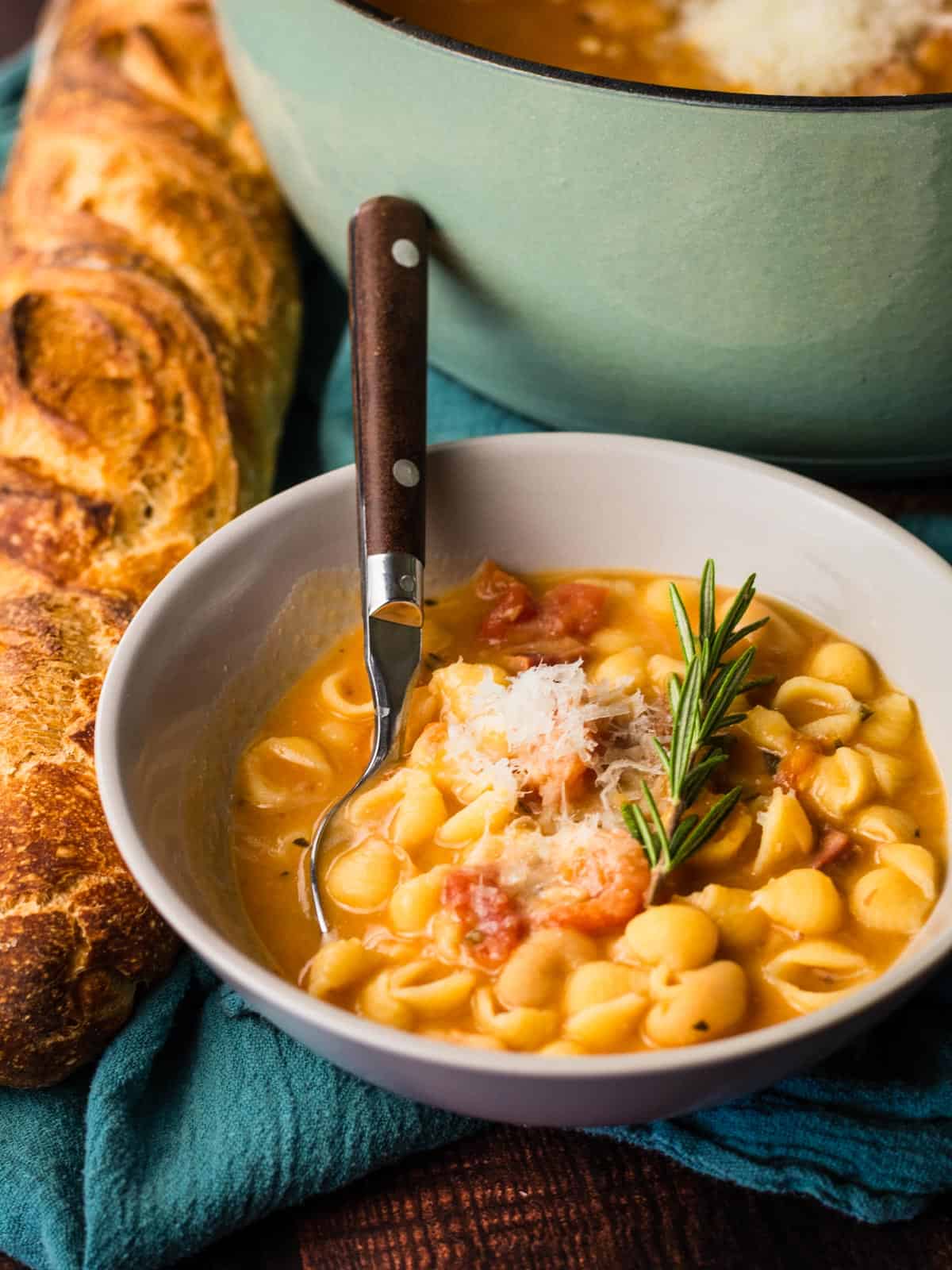 bowl of pasta fagioli with a spoon in it and rosemary and freshly grated cheese on top.