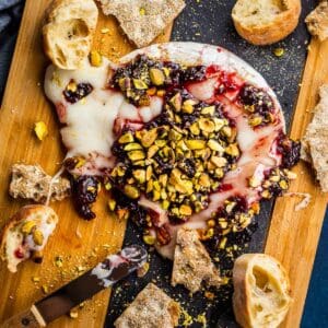 baked brie melted on a cutting board topped with jam and pistachios with bread around.