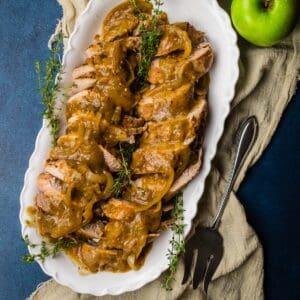 sliced pork tenderloin topped with gravy on a white platter with fresh thyme and green apples.