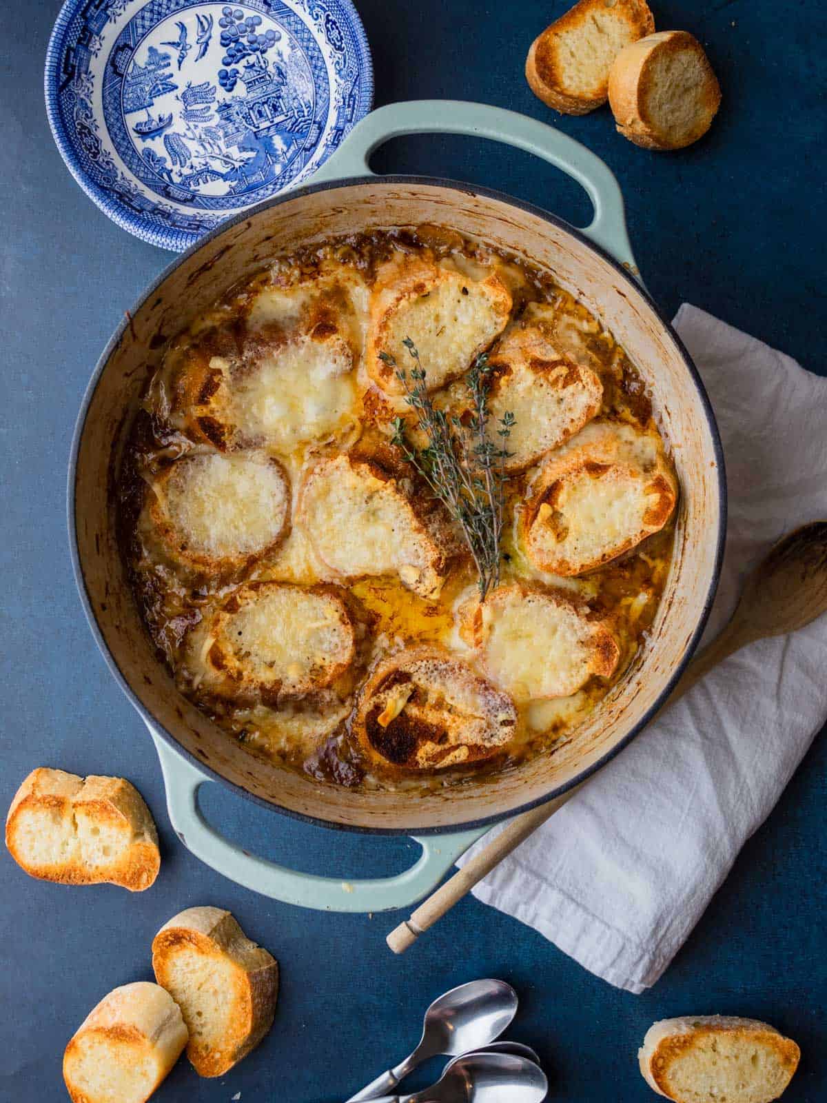 french onion soup in dutch oven with bowls, spoons and toasted croutons lying around.