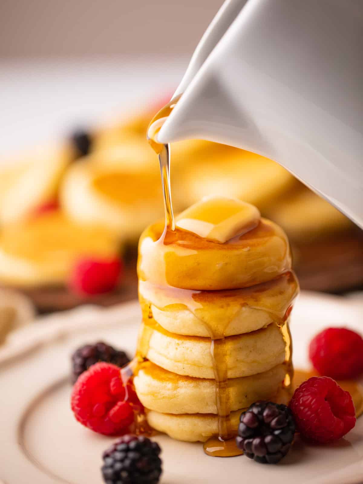 syrup pouring over a stack of mini pancakes with butter on top and fresh berries around.
