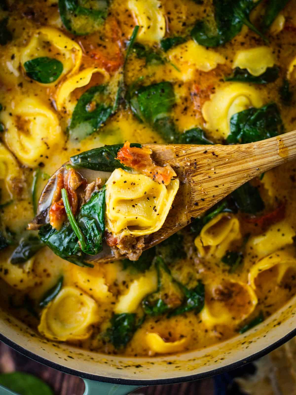 wooden spoon holding a scoop of tortellini soup with spinach and sundried tomatoes.