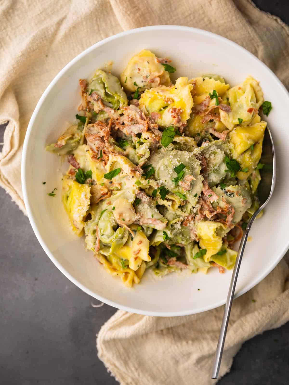 white bowl filled with green and yellow tortellini in a cream sauce with prosciutto.
