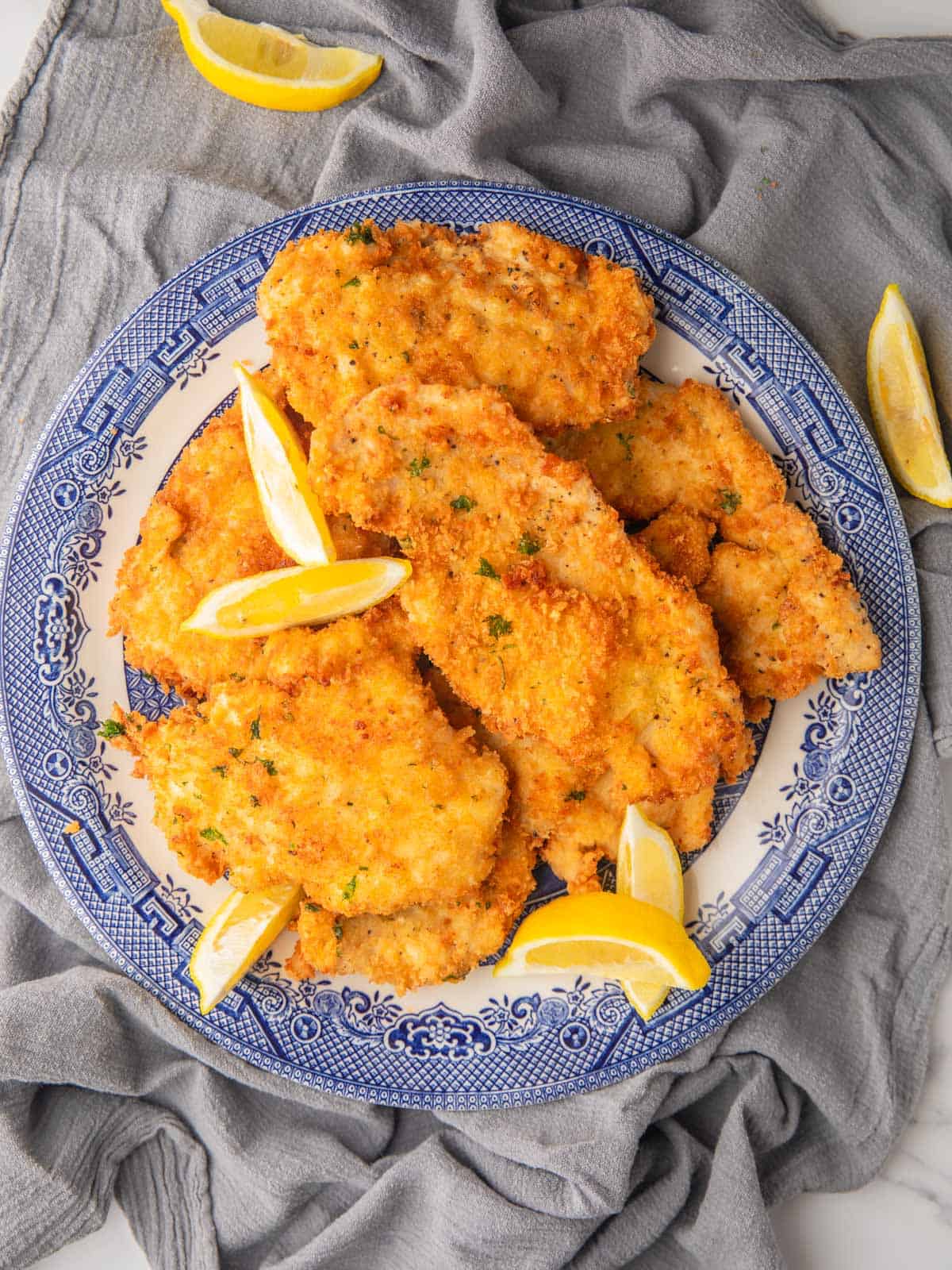 crispy chicken cutlets on a round blue and white plate with lemon wedges around.