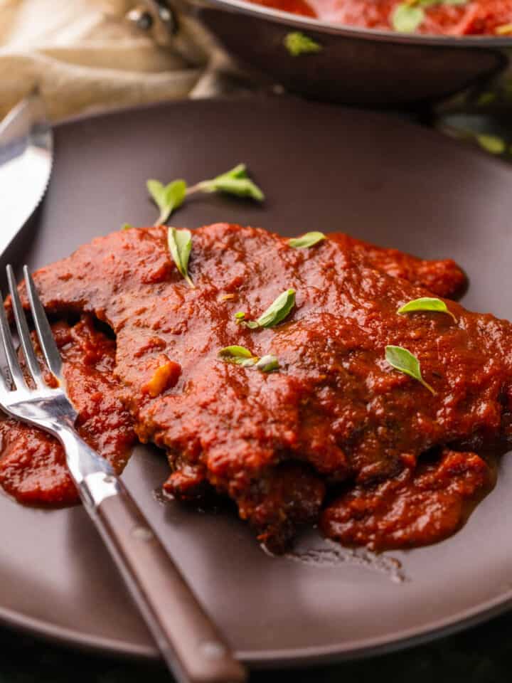 steak covered in red sauce on brown plate with fresh oregano, a fork and steak knife.